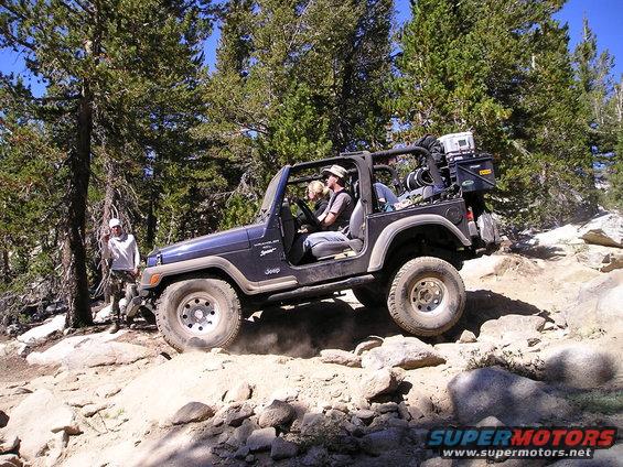 Dark blue Jeep Wrangler TJ off-roading on a rocky trail with two people inside and another person standing nearby