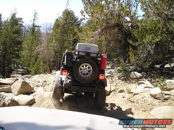 Jeep Wrangler driving over rocks on a dirt trail, spare tire on back, cooler on roof rack