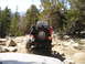 1989 Ford Bronco - A Jeep navigates a rocky off-road trail with trees and boulders surrounding it.