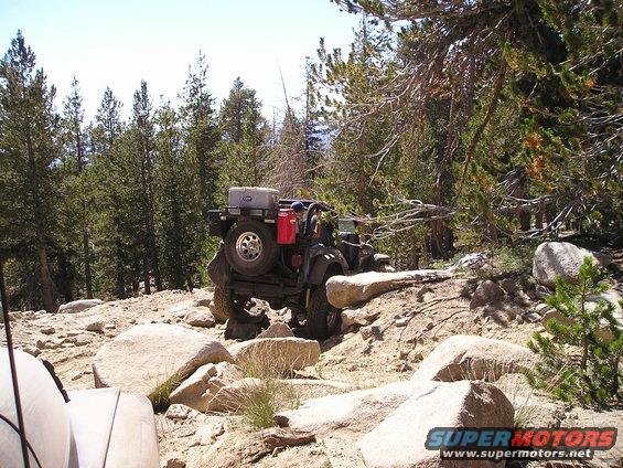 Black off-road vehicle with spare tire and gas cans on rear, navigating rocky terrain