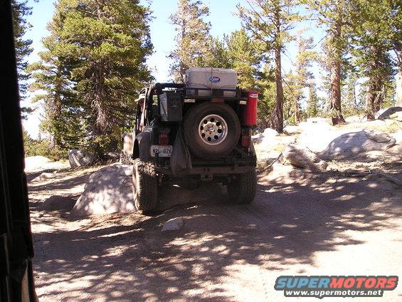 Rear view of a Jeep Wrangler with a spare tire, cooler, and red canister on the back, driving on a dirt trail