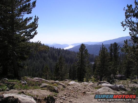 Mountain landscape with pine trees, rocks, and a distant lake under a clear blue sky