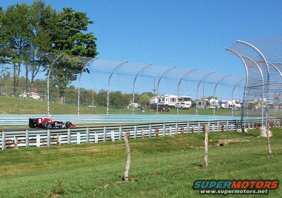 Race car with red and black livery on a track, with RVs parked in the background