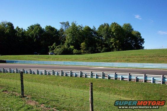 Two cars are parked on a grassy hill next to a racetrack with blue and white barriers