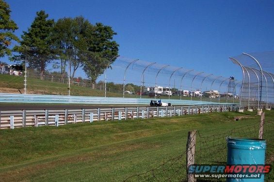 Open-wheel race car on a track with RVs and trees in the background