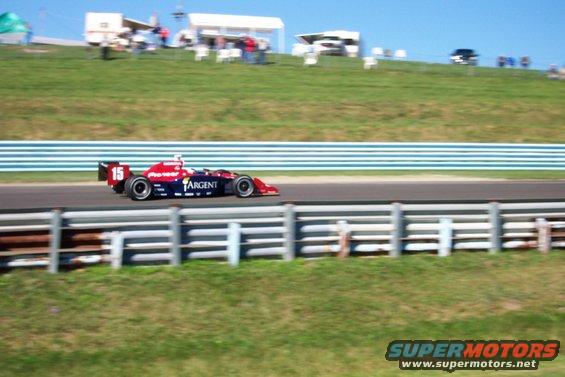 Red and blue open-wheel race car with number 15 on the rear wing, moving on a track
