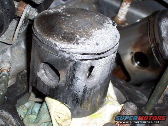 Damaged piston with white deposits and a hole in its side, next to another piston