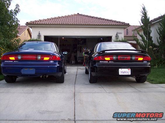 two 1995 Ford Probes parked in a driveway, one blue and one black, facing a garage