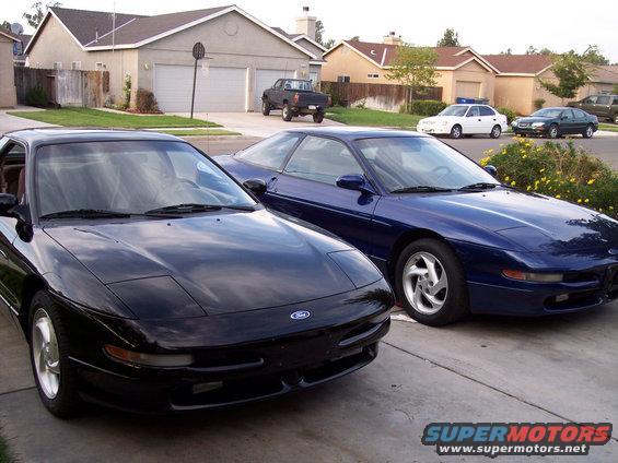 Two Ford Probes parked in a driveway, one black and one blue. Houses and a pickup truck are in the background