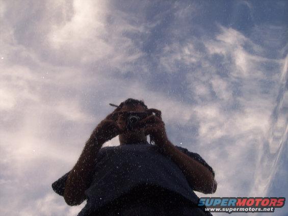 Reflection of a person holding a camera, against a cloudy sky, on a car's surface