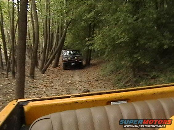 Black 1986 Ford Bronco driving on dirt road through trees