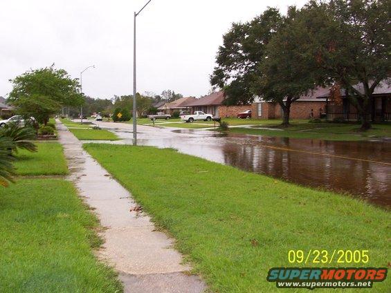 Flooded street in a residential area with houses and trees visible. A white pickup truck and a red car are parked in driveways