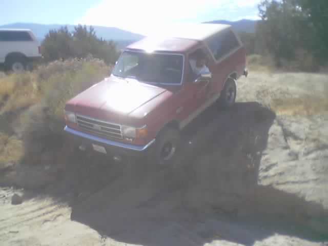 Red 1990 Ford Bronco driving off-road, with another white SUV in the background