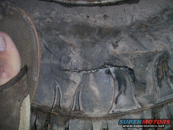 Close-up of a damaged tire tread with a visible cut and wear. A person's thumb is visible on the left