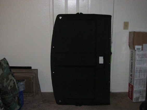 Black car headliner panel leaning against a wall, with boxes and camouflage fabric nearby