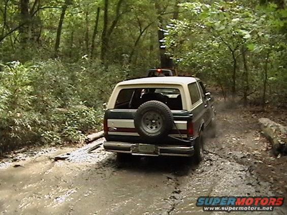Dark green and tan Ford Bronco driving through mud, spare tire on rear door