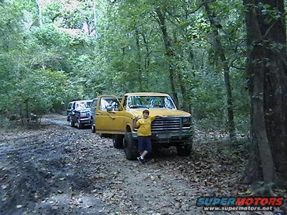 Yellow 1986 Ford Bronco with open door, person leaning on hood, forest road