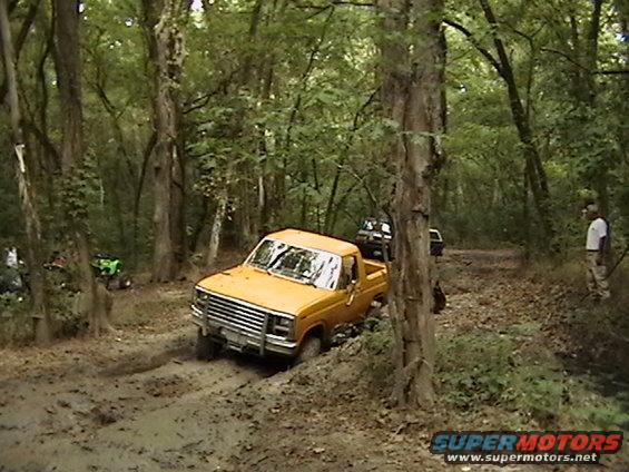Yellow pickup truck driving through mud on a wooded trail. Another truck and an ATV are visible in the background