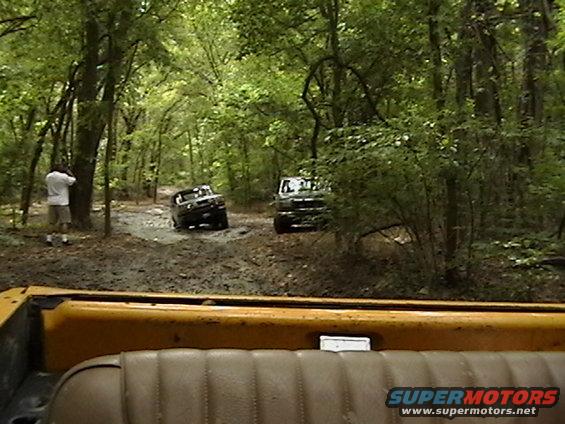 two SUVs driving on a muddy forest road, one in front of the other, with a person standing by a tree