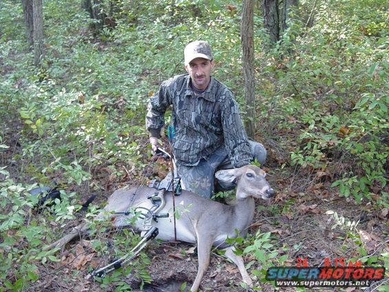 Hunter kneels next to a harvested deer in a wooded area, holding a bow