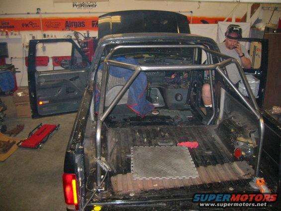 Truck interior with a rollcage installed, doors open, hood up. Person on phone in driver's seat