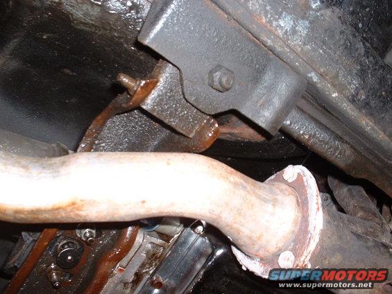 Exhaust pipe flange with white sealant and rusty bolts, attached to a black metal bracket