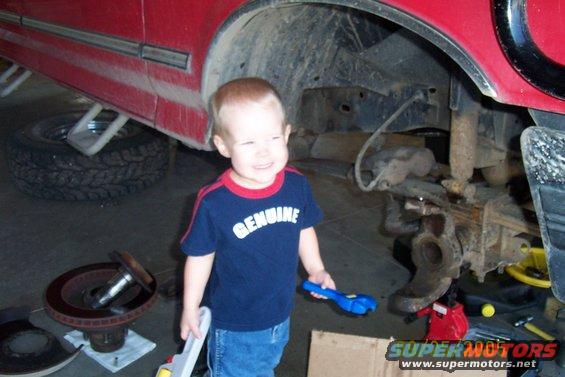 Kid in garage holding toy tools near disassembled truck suspension and brake rotor