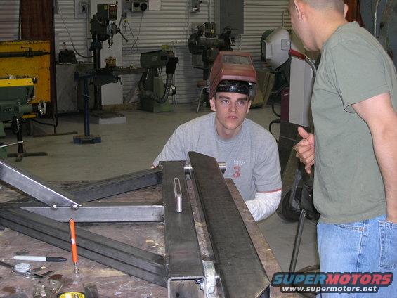 Two men in a workshop, one wearing a welding mask, looking at metal parts on a workbench