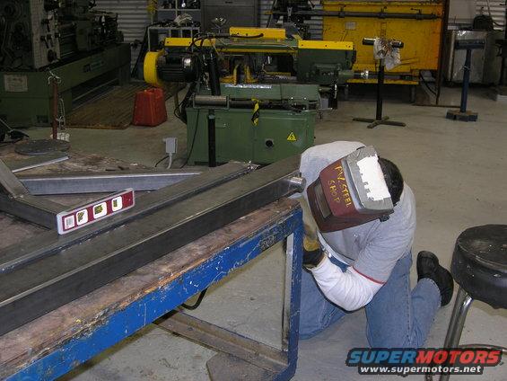 Guy in welding mask working on metal frame on a workbench in a shop. Level on the metal