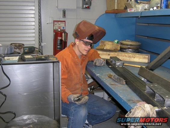 Welder wearing a mask and safety glasses, leaning on a workbench with metal parts