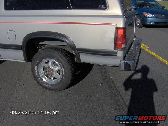 Pickup truck rear wheel well, bumper, and taillight; blue sedan in background