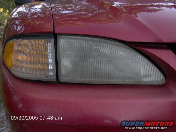 Close-up of a red car's front passenger side headlight and amber turn signal. The headlight lens is ribbed