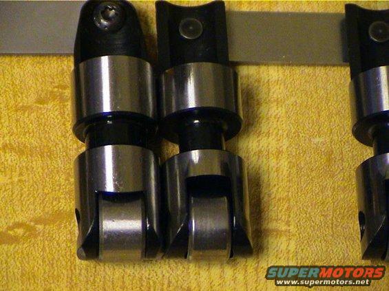 Engine lifters with metal rollers and black tops, arranged in a row on a yellow textured surface