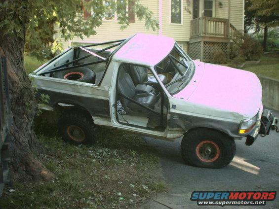 White and grey Ford Bronco with roll cage, off-road tires, and spare tire in bed, parked on grassy incline