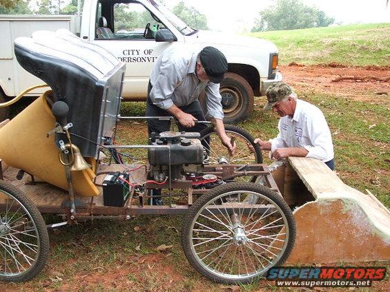 Two men working on a custom-built vehicle with bicycle wheels and a small engine. A white pickup truck is parked behind them