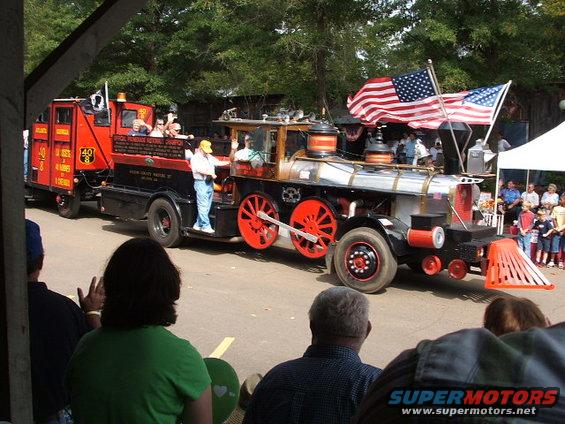 Custom locomotive vehicle with a fire truck trailer, decorated with American flags, in a parade
