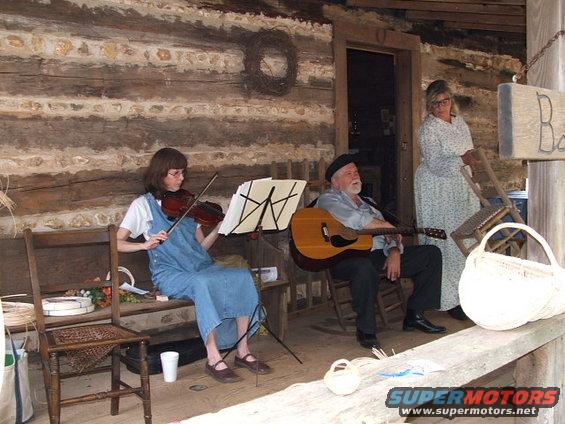 People playing music on a porch with a log cabin background