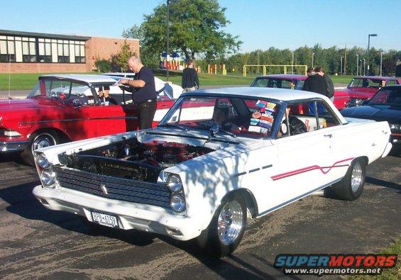 White classic coupe with hood open, parked next to a red car. Some stickers on the windshield