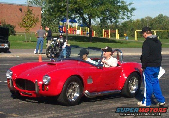 Red convertible sports car with a roll bar, two people inside, parked on asphalt