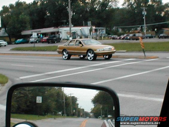 Gold sedan with large chrome wheels driving on a road, seen from a side mirror