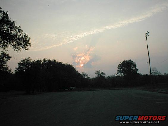 Empty asphalt parking lot with trees and a light pole against a cloudy sky at dusk
