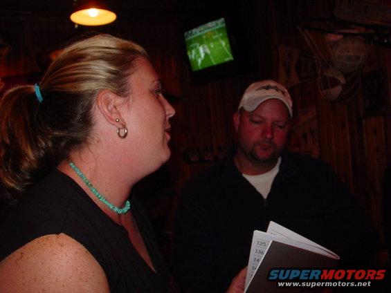 Two people, a woman and a man, are seated at a table. A TV displays a football game