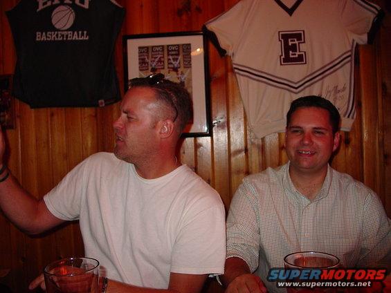 Two men sit at a table in a wood-paneled room with sports memorabilia on the wall