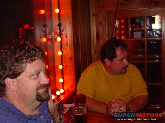 Two guys sitting at a bar with orange string lights behind them. One man wears a blue shirt, the other a yellow shirt