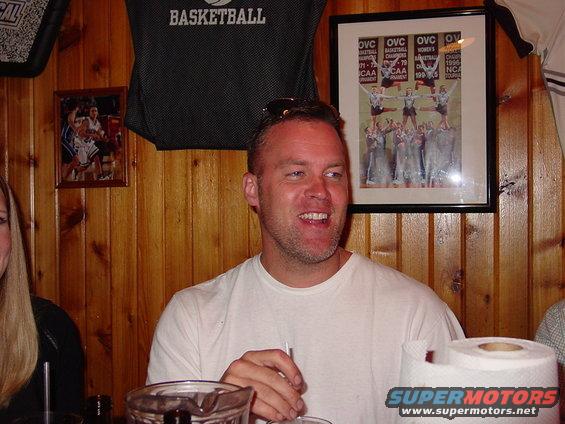 Man in white t-shirt smiling, with framed photos and a basketball shirt on a wood-paneled wall