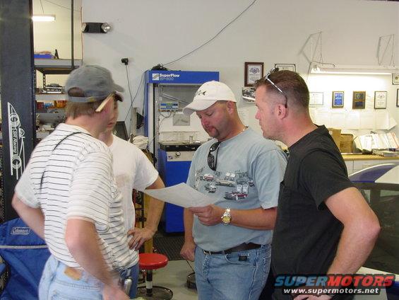 Four men gathered around a document in a garage setting with a car lift and equipment visible