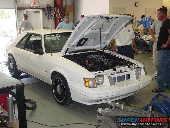 white 1986 Ford Mustang coupe, hood open showing engine, people around in garage