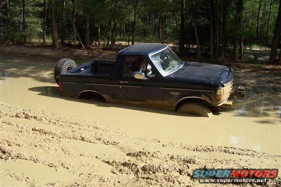Black 1987 Ford Bronco stuck in mud, spare tire visible on rear, trees in background