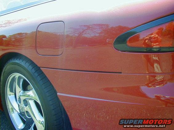 red car's rear quarter panel, gas cap door, and taillight. Reflections of trees and a person are visible