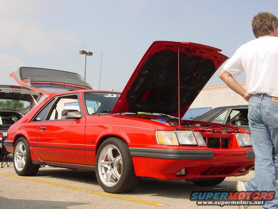 Red coupe with hood and trunk open, parked next to another car. Guy in jeans and white shirt stands nearby